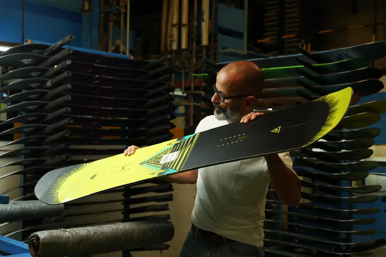 A man inspecting a black-and-yellow snowboard in front of racks of boards at a factory.